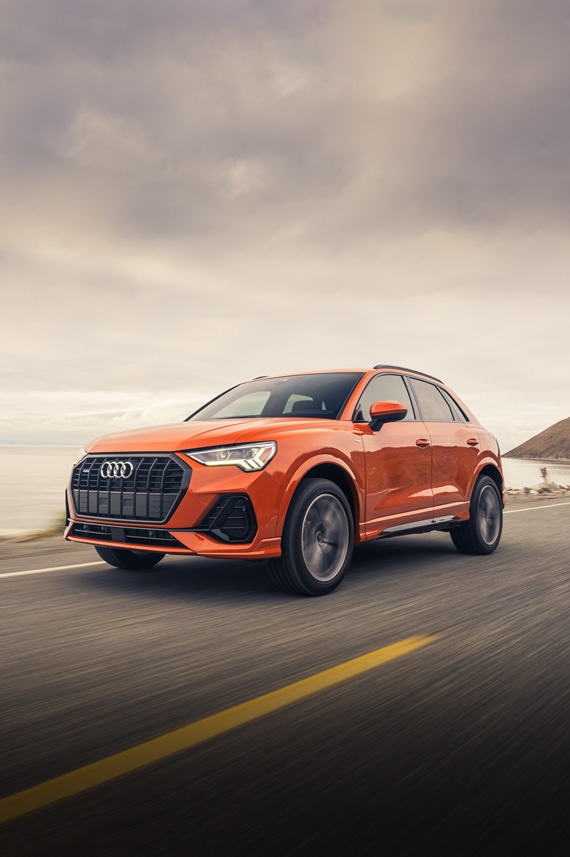Three-quarter front view of an orange Audi Q3 accelerating on an oceanside road.