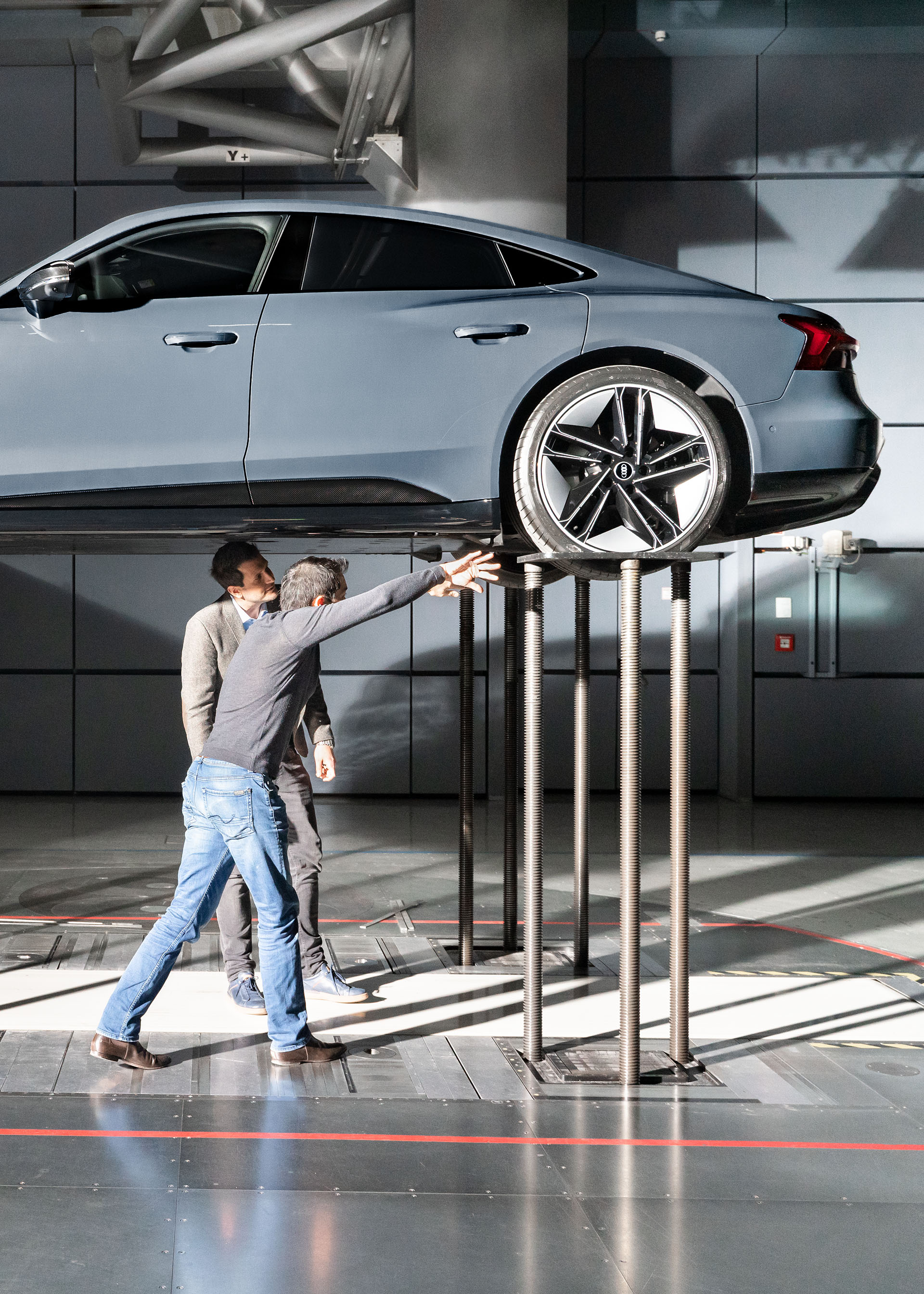 Two Audi aerodynamicists inspect the underbody of an Audi RS e-tron GT on a raised platform. 