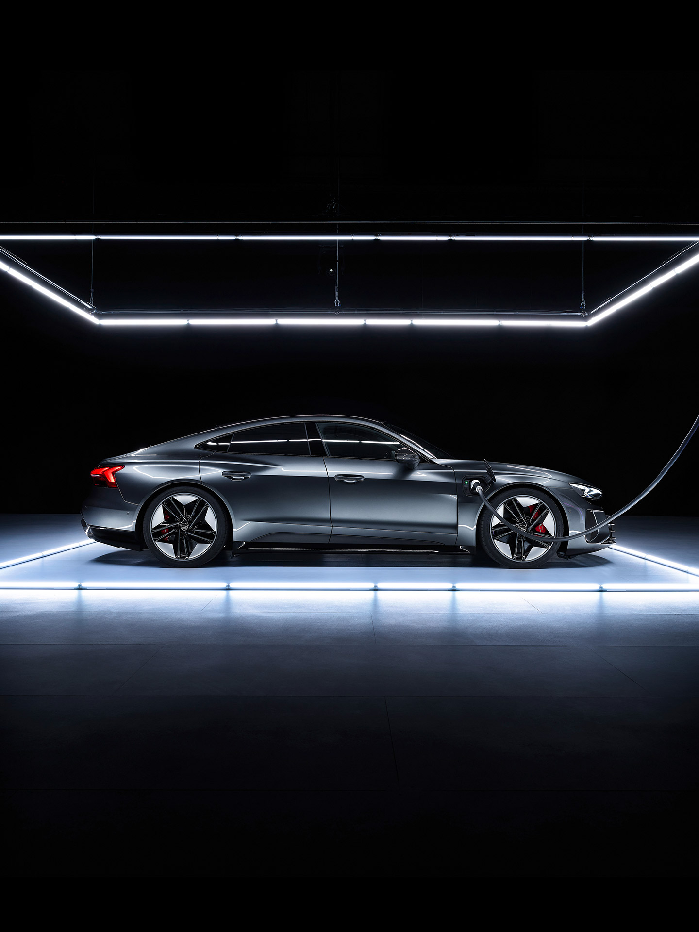 Shifting image of a grey Audi e-tron GT parked in front of a daytime city skline and on an illuminated platform in a dark room. 